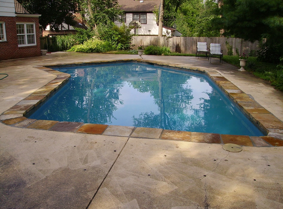 A tranquil backyard pool is framed by stone tiles, reflecting the nearby trees and sky. Patio chairs and lush greenery surround the serene setting.