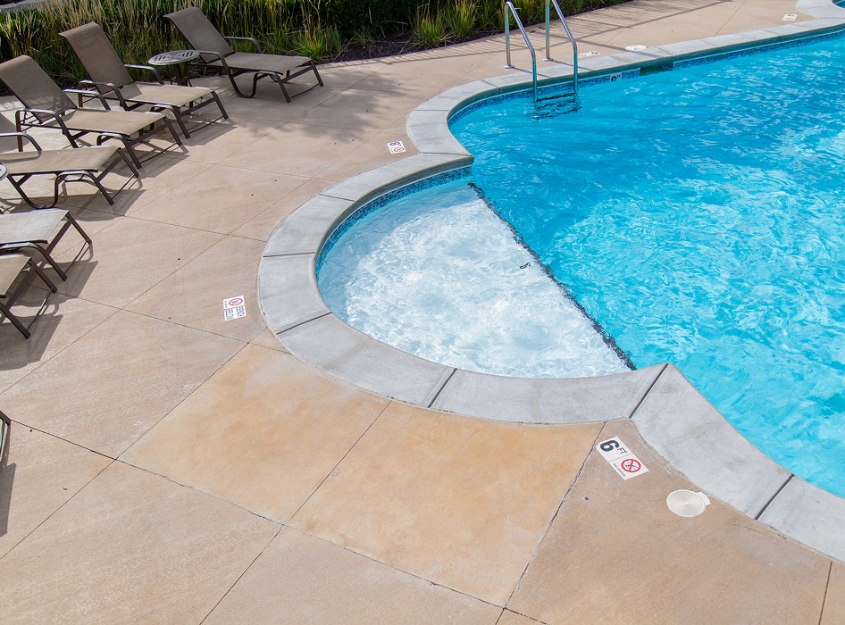 A curved swimming pool with a bubbling jacuzzi section and metal steps at the side. Nearby are several beige lounge chairs on a concrete patio. The setting is calm and inviting.