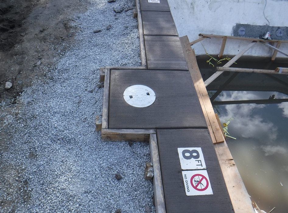 A concrete pool edge under construction with wooden forms and gravel. A sign reads "8 FT, No Diving" near calm water reflecting the cloudy sky.