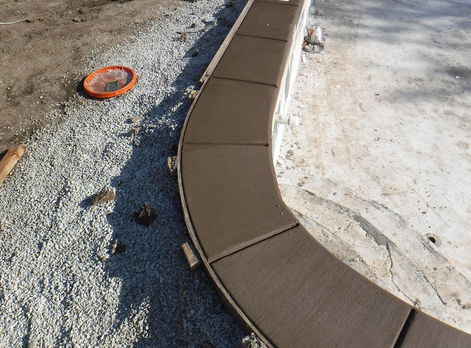 Freshly poured curved concrete edge around a pool, surrounded by gravel. The wet surface shines in the sunlight, conveying a work-in-progress feel.