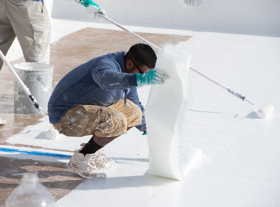 A person wearing a blue hoodie and gloves kneels on a white surface, laying fiberglass matting, while another uses a roller to apply coating. Industrial setting.