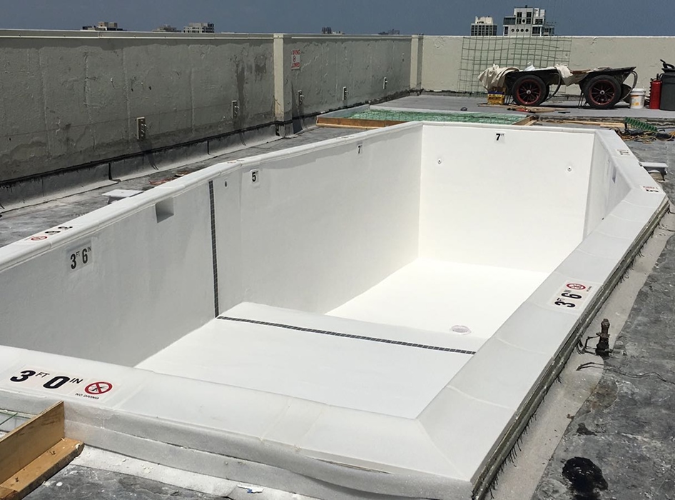 Empty rooftop swimming pool under construction, with white interior and depth markers "3' 0''" and "3' 6''." Background shows urban skyline.
