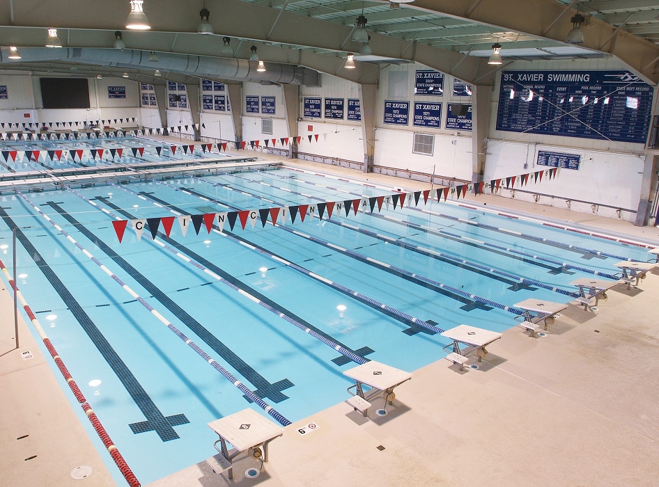 Indoor swimming pool with multiple lanes, clear water, and red-and-white lane markers. Diving platforms line one side. Bright, well-lit atmosphere.