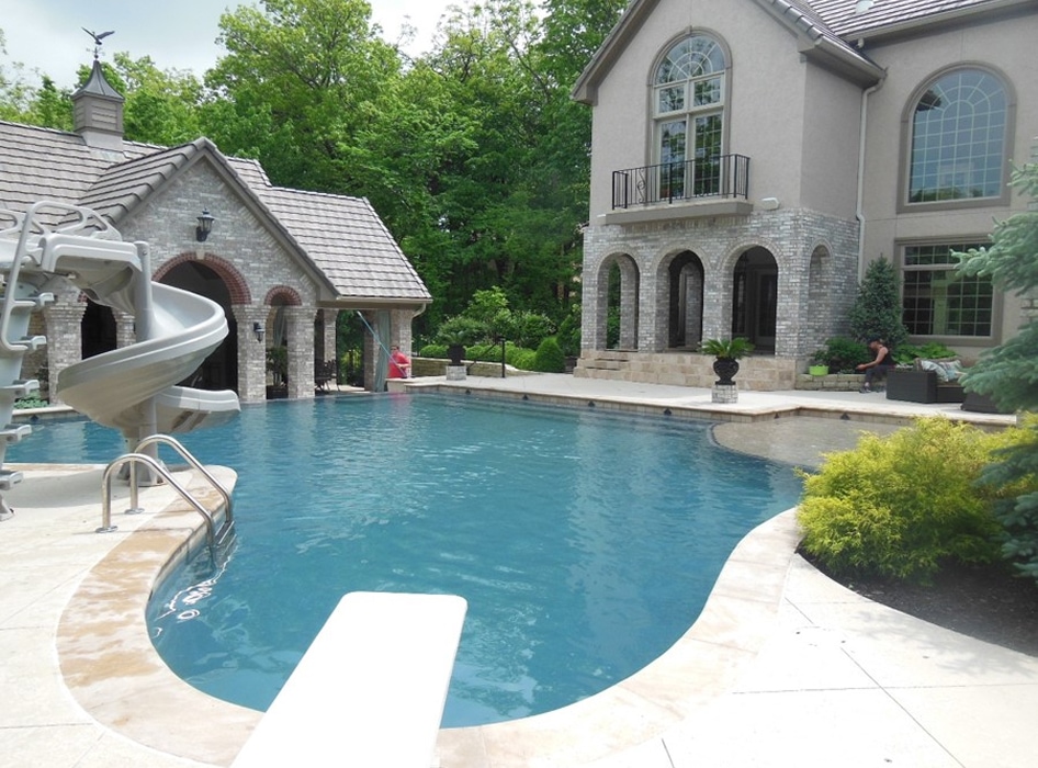 Luxurious backyard with a swimming pool featuring a slide and diving board, surrounded by a stone patio, elegant house, and lush greenery.