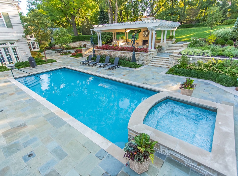 Luxurious patio scene with a crystal-clear pool and adjacent hot tub. Elegant stone tiles, lounge chairs, a gazebo, and lush greenery create a serene, upscale atmosphere.