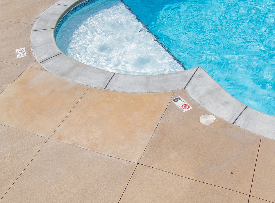Curved pool corner with clear blue water against beige pavement. Depth marker reads 6 feet, surrounded by no diving symbols. Bright, sunny setting.