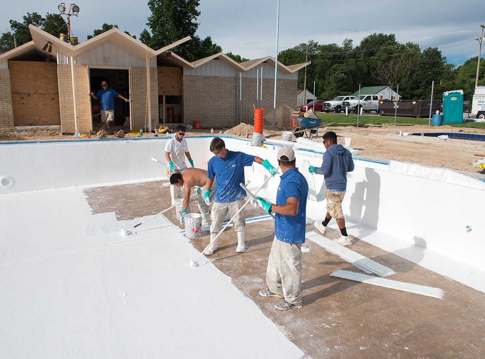 Five workers paint the interior of an empty pool white on a sunny day, with tools and materials scattered around. A partially constructed building is visible in the background.