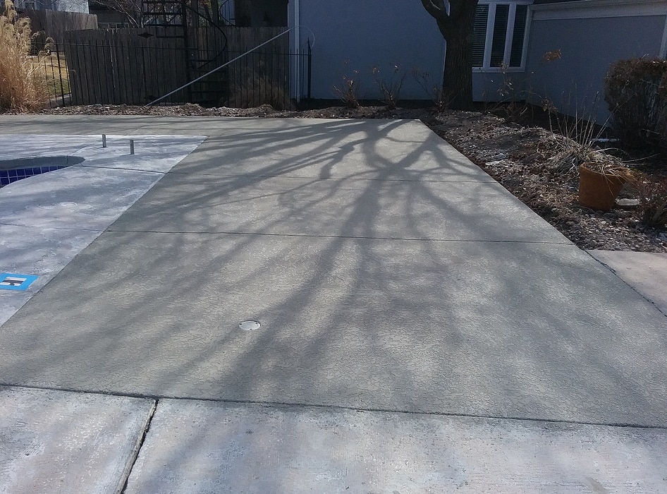 Concrete patio with bare tree shadows cast across it, beside a closed pool. A dark fence and winter plants surround the area, creating a quiet, somber tone.