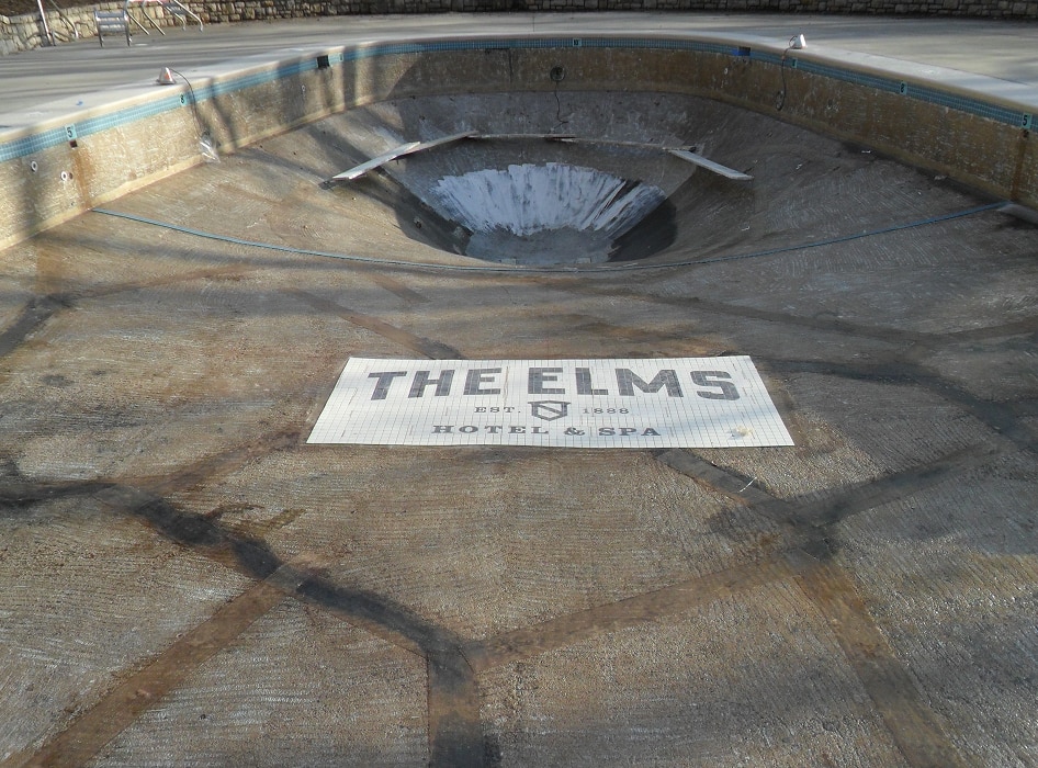 An empty, cracked swimming pool with a sloped center and a mosaic sign on the floor reading "The Elms Hotel & Spa" conveys a sense of abandonment.