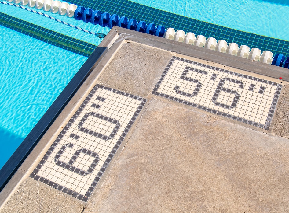 Close-up of a swimming pool edge showing depth markers "6.0" and "5'6" against a bright blue water backdrop, featuring lane dividers. Sunny day ambiance.