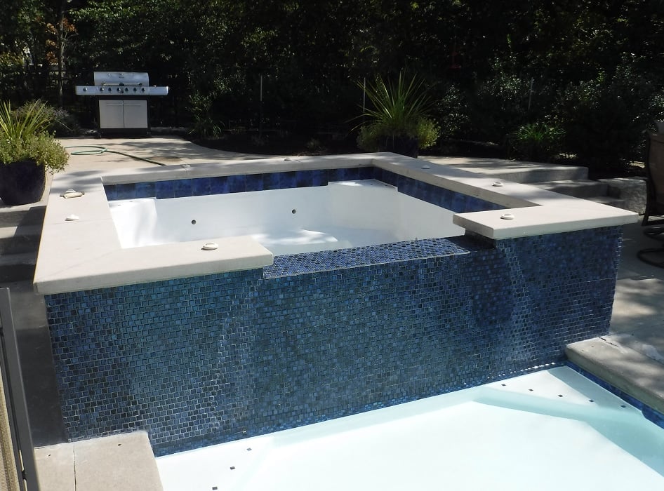 Outdoor scene of an empty square hot tub with blue mosaic tiles adjacent to a small pool. Background features a patio, grill, and lush greenery.