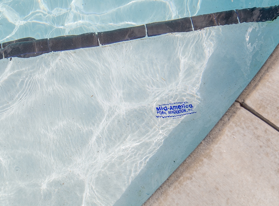 Close-up of a pool corner with rippling water, showing a tiled edge and a logo on the pool floor reading "Mid-America Pool Renovation, Inc." Calm and clean.