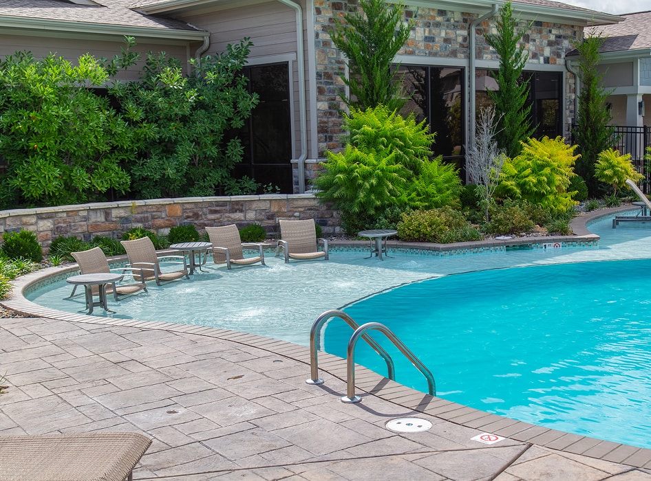 Outdoor pool area with turquoise water, surrounded by lounge chairs and tables on a tiled deck. Lush greenery and stone walls enhance the serene setting.