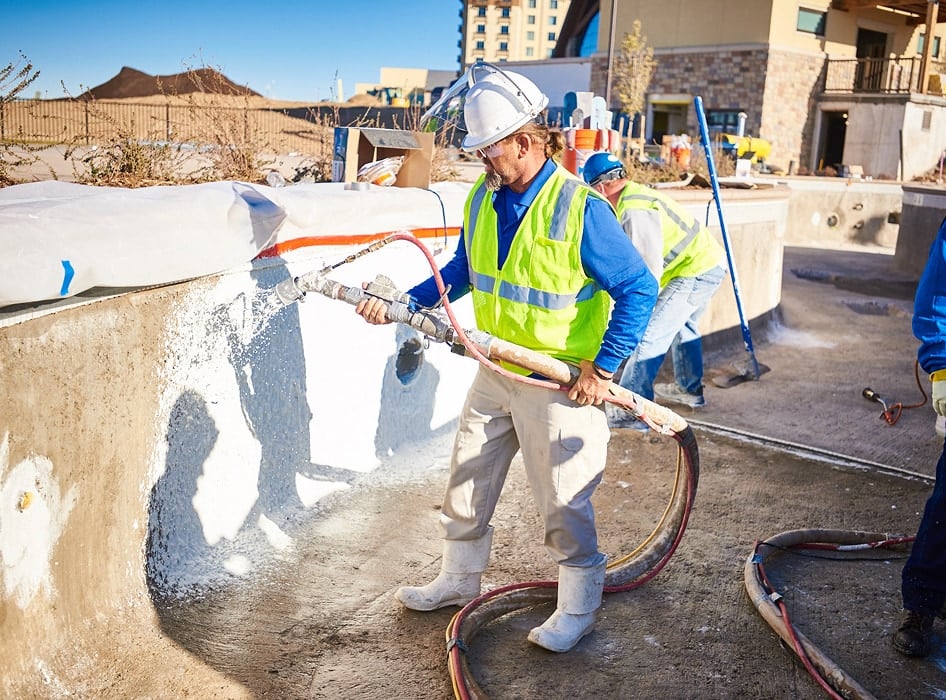 Worker in a hard hat and safety vest applies concrete to a pool edge with a hose, shadow cast on surface. Sunny day, construction site in background.