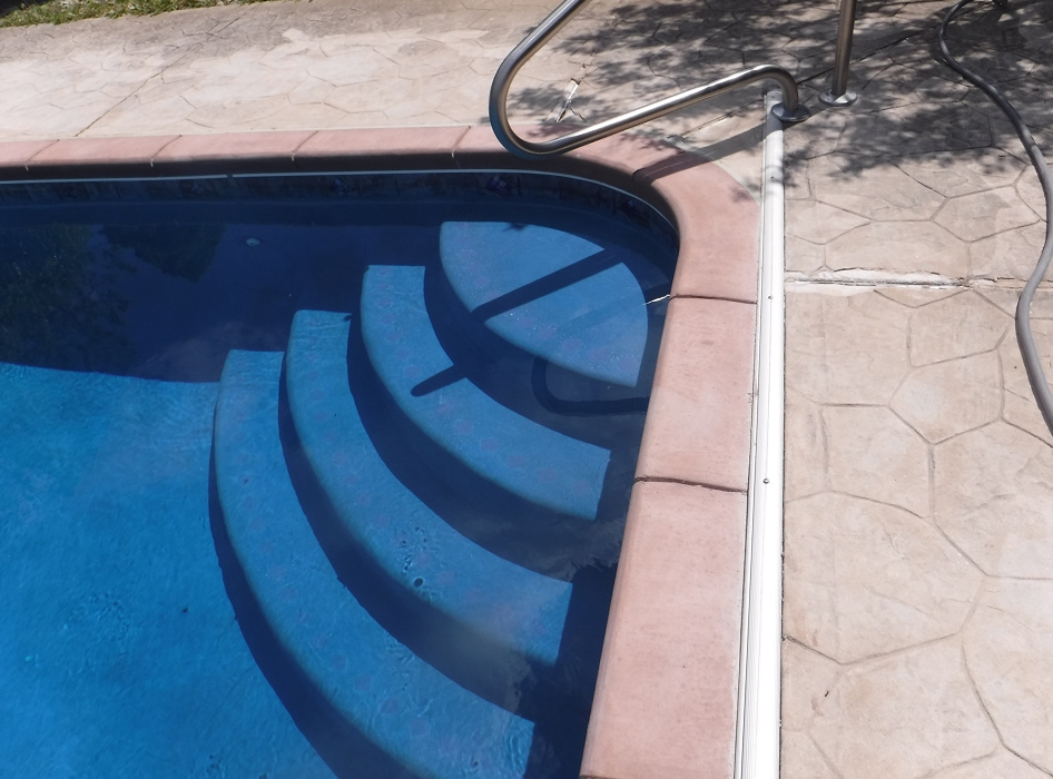 Curved pool steps descend into a clear blue swimming pool, bordered by pink tiles. Sunlit concrete surrounds the area, creating a serene, inviting atmosphere.