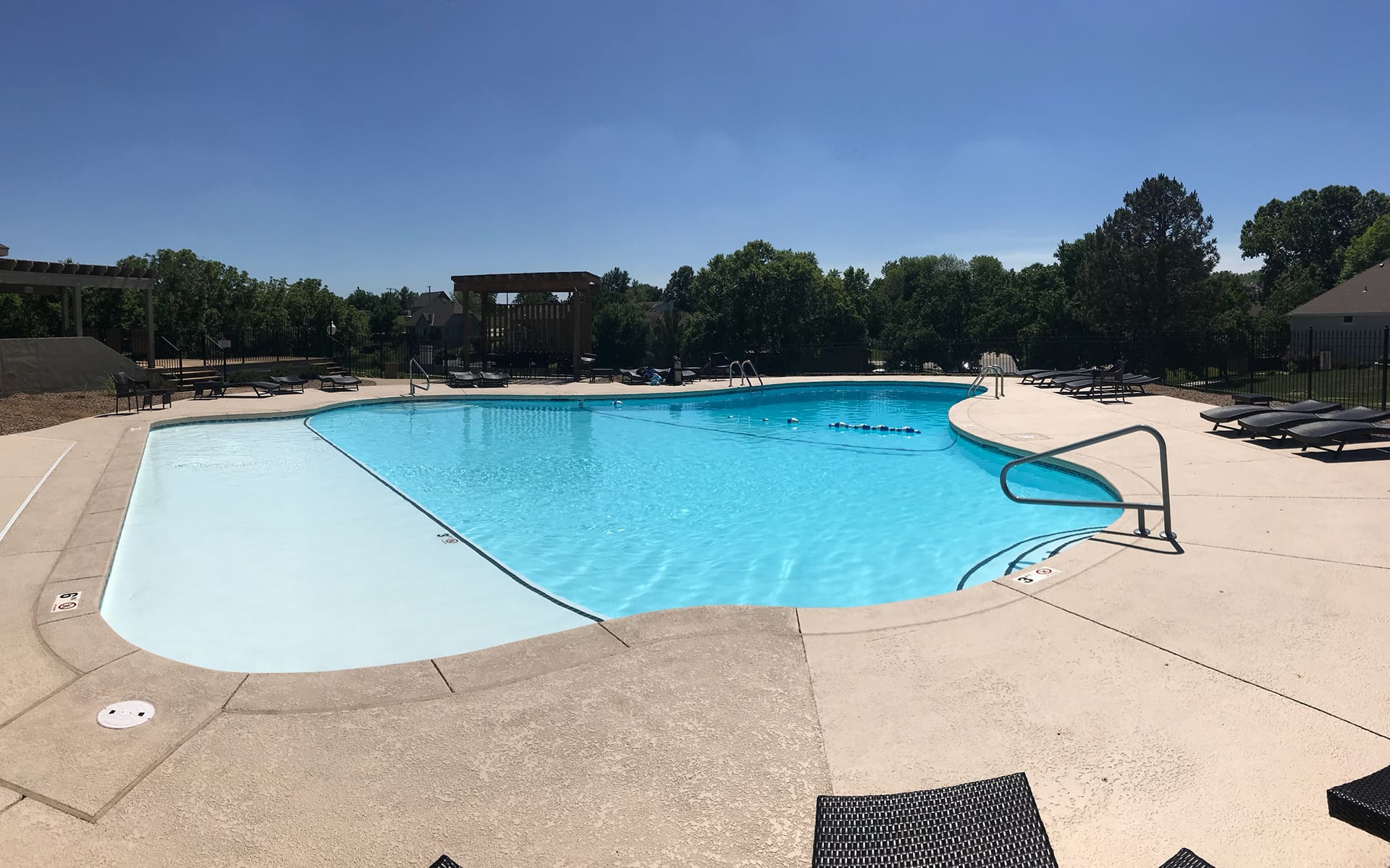 Large outdoor swimming pool under a clear blue sky, surrounded by lounge chairs and trees. The scene is peaceful and inviting, evoking relaxation.