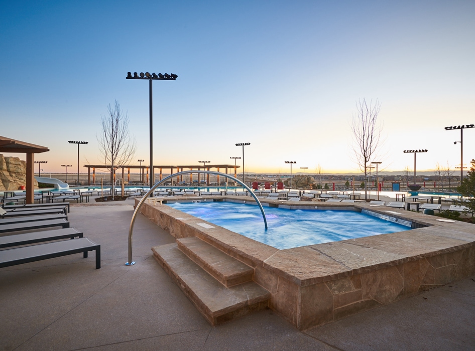 Outdoor pool area at sunset with a square hot tub and cascading water feature. Surrounding the pool are lounge chairs, bare trees, and distant hill views. The scene is tranquil and inviting.