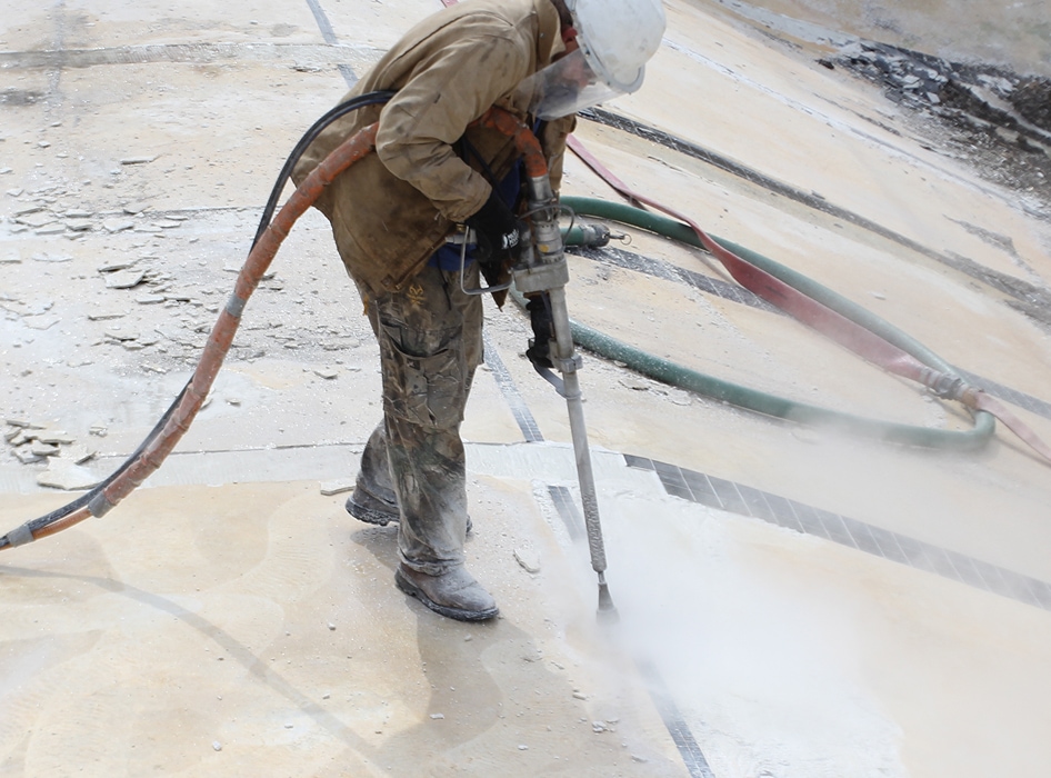 A worker in protective gear uses a high-pressure water jet to remove surface layers from concrete. The environment appears industrial and dusty.
