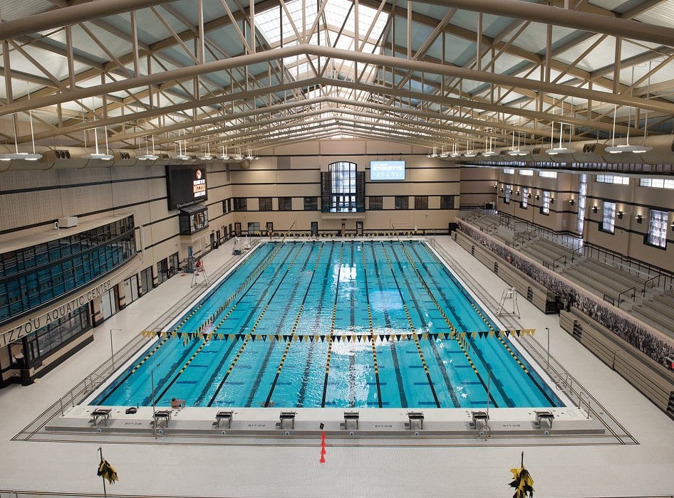 Indoor swimming pool with multiple lanes, enclosed by bleachers on the right and a wall of windows on the left. The space is lit by overhead lights and natural light through the roof, creating a bright and spacious atmosphere.