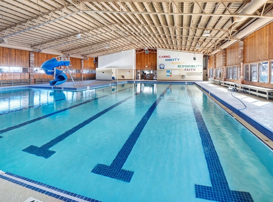Indoor swimming pool with clear blue water and lane markings. A blue slide is on the left, and a wall displays the words "Caring, Honesty, Responsibility, Faith." Warm lighting and wood paneling create a welcoming atmosphere.