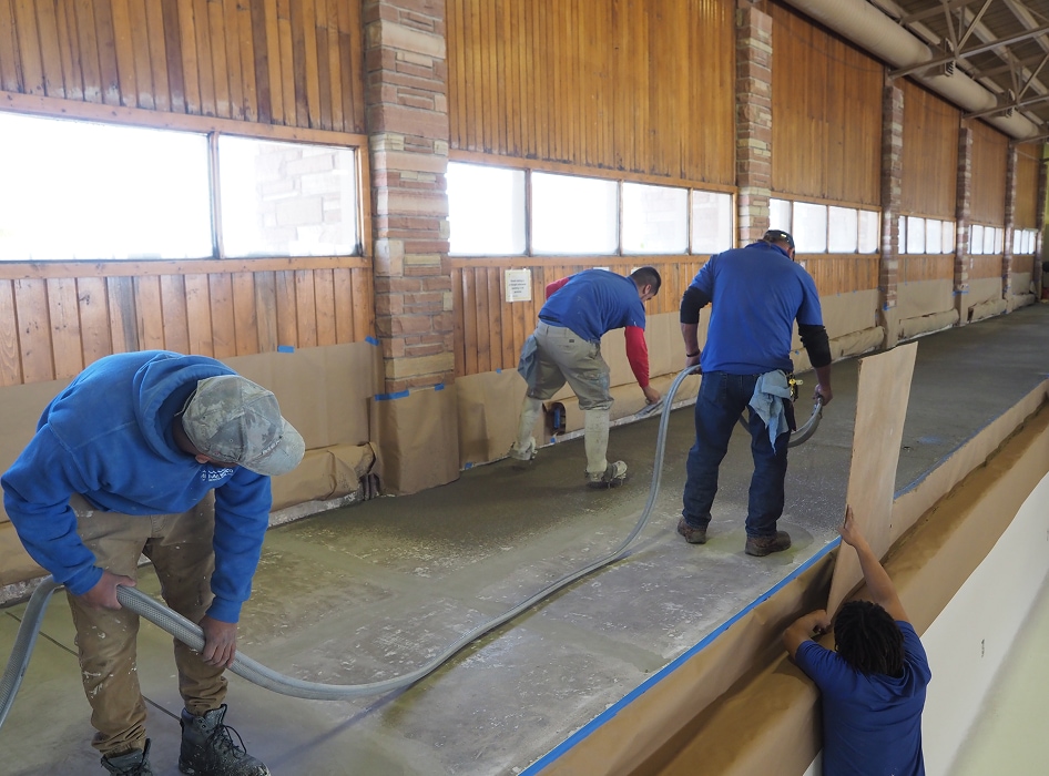 Four workers in blue shirts are smoothing concrete on a large indoor floor. The room has wood-paneled walls and a focused, industrious atmosphere.