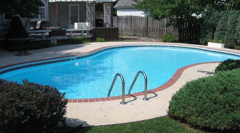 Residential freeform swimming pool with clear blue water red brick border concrete deck and backyard patio furniture