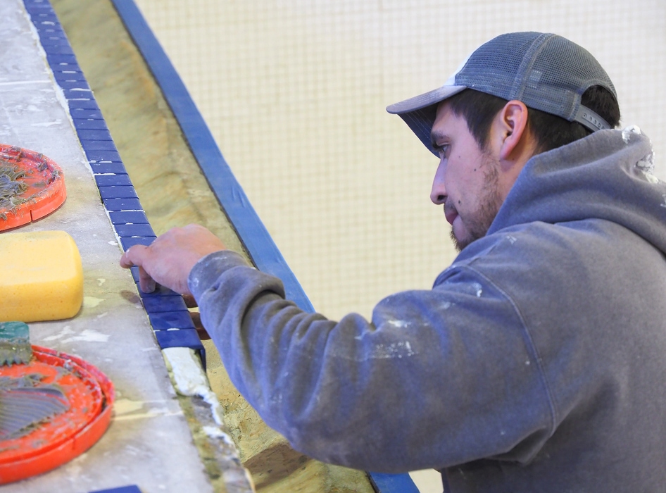 A man wearing a baseball cap and hoodie carefully aligns small blue tiles on a surface. Nearby are sponges and tools, suggesting a calm and focused mood.
