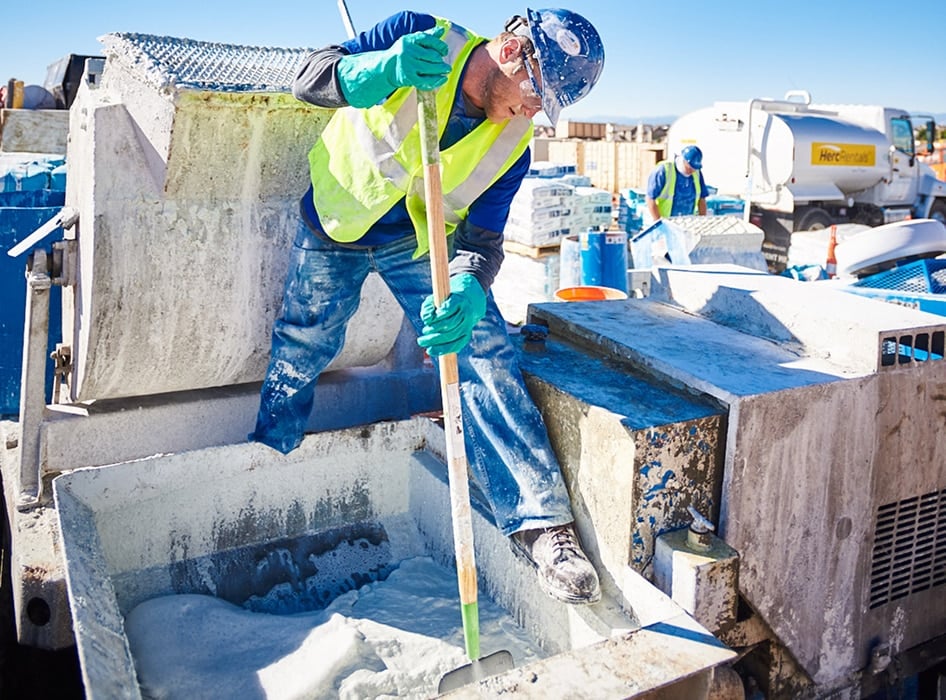 A construction worker in a safety vest and helmet stirs white cement in a mixer using a long stick. The outdoor scene is bright and busy, with another worker and equipment in the background. The atmosphere is industrious and focused.