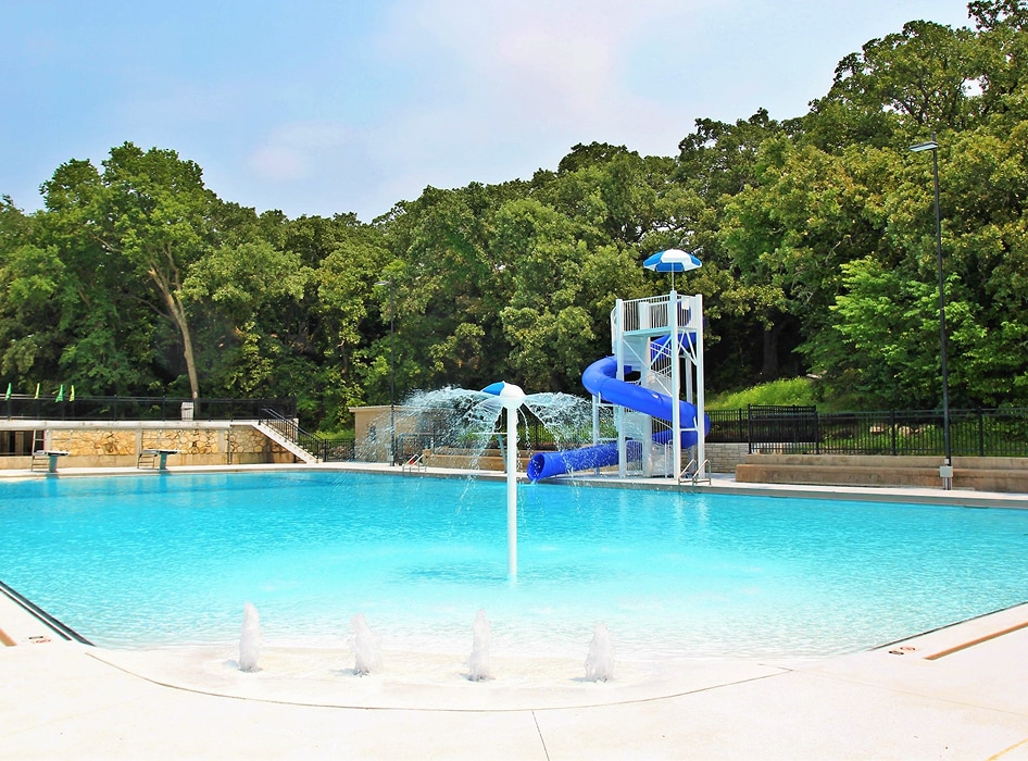 A large outdoor swimming pool with clear blue water, features a playful blue water slide and fountains, surrounded by lush green trees under a clear sky.