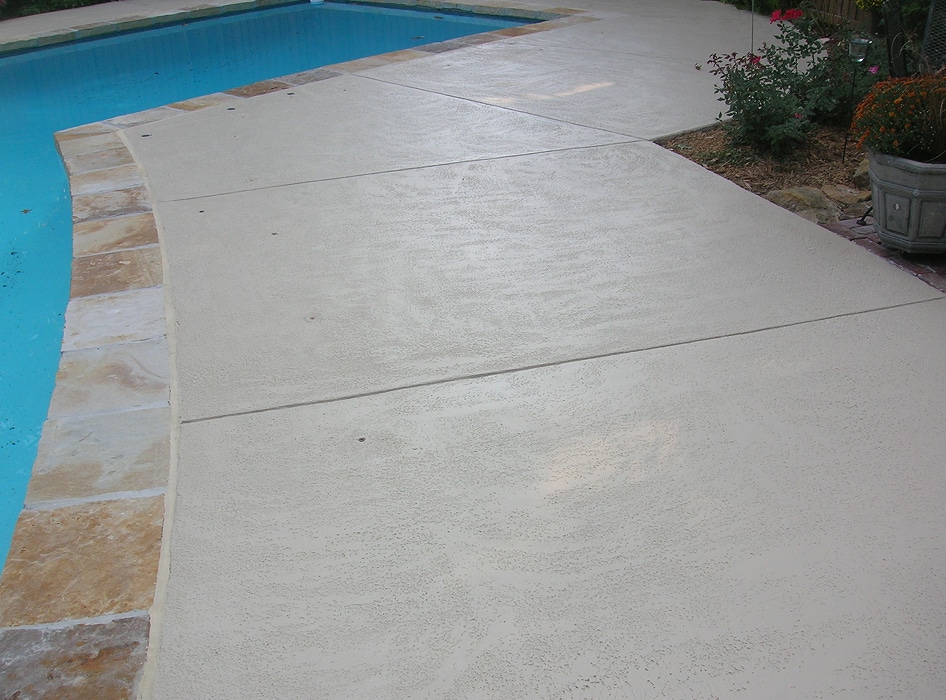 Smooth, light beige textured concrete pool deck with stone edging borders a blue pool. Nearby, potted plants and shrubs add greenery.