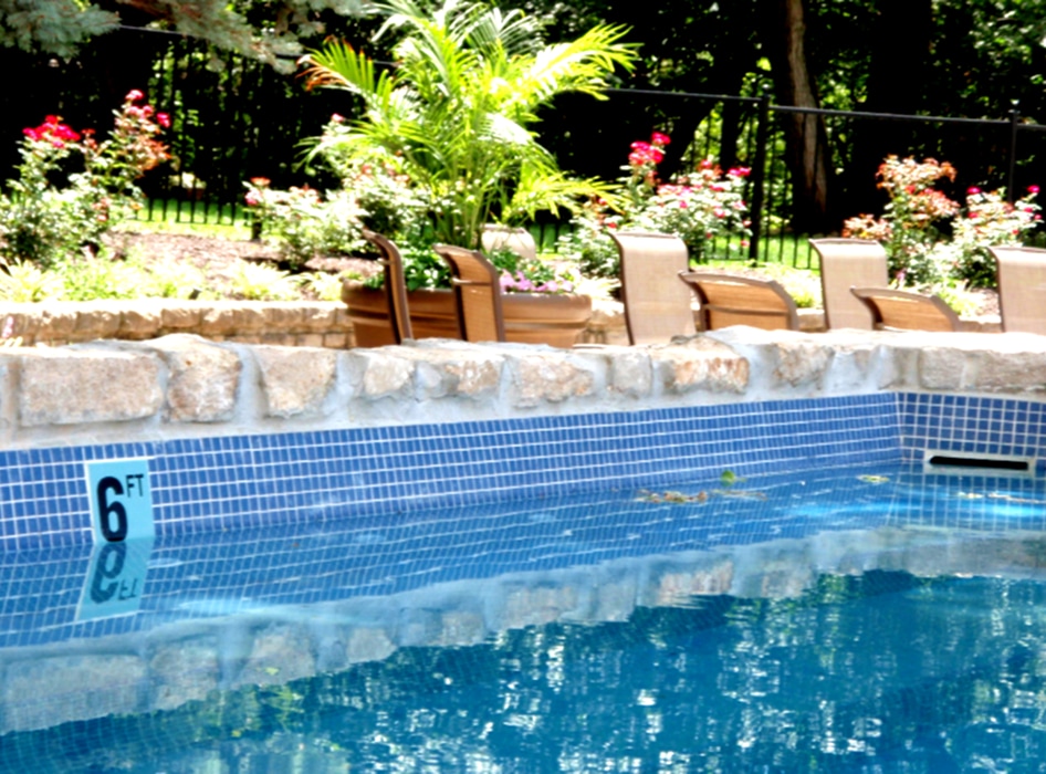 Outdoor pool with blue tiles marked at 6 feet, surrounded by stone ledge. In the background, patio chairs and lush greenery create a serene atmosphere.