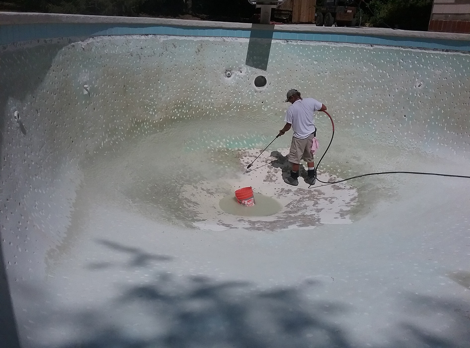 A person cleaning an empty, worn swimming pool with a pressure washer. The scene is sunny, with visible shadows, and there's a red bucket nearby.
