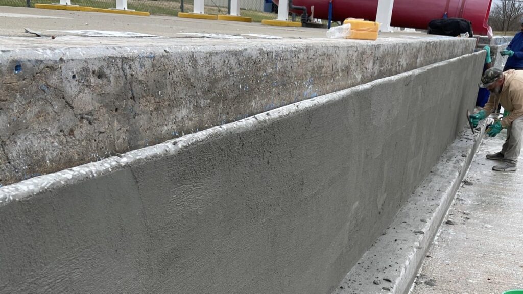 Man wearing gloves applies plaster to a large concrete wall, creating a smooth surface. The scene conveys a sense of manual labor and construction.