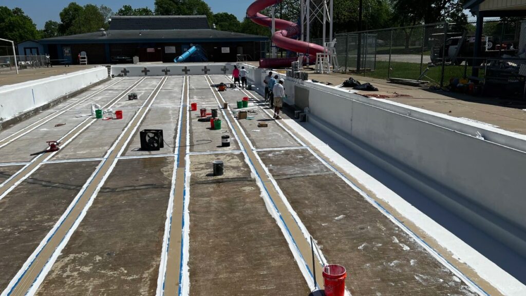 An empty outdoor pool under renovation with workers painting the floor. Paint cans and tools are scattered around. A large red slide is in the background.