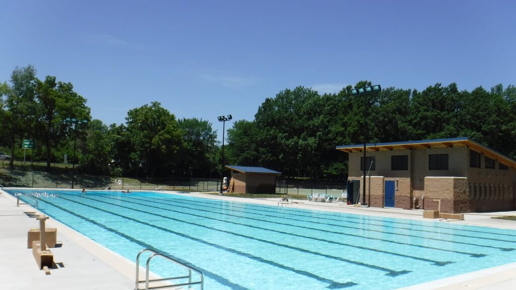 Outdoor swimming pool under a clear blue sky, surrounded by trees. A pool building with a sloped roof is on the right. The scene is calm and inviting.