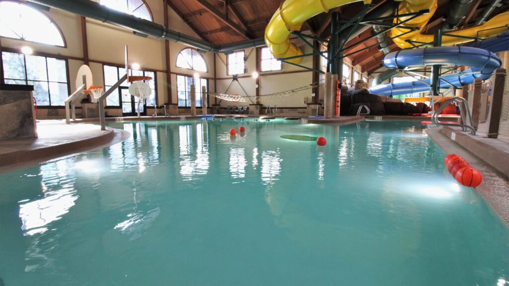 Indoor water park with a large pool, illuminated by natural light from arched windows. Bright yellow and blue slides curve overhead, and a basketball hoop is on the left. The atmosphere is playful and inviting.
