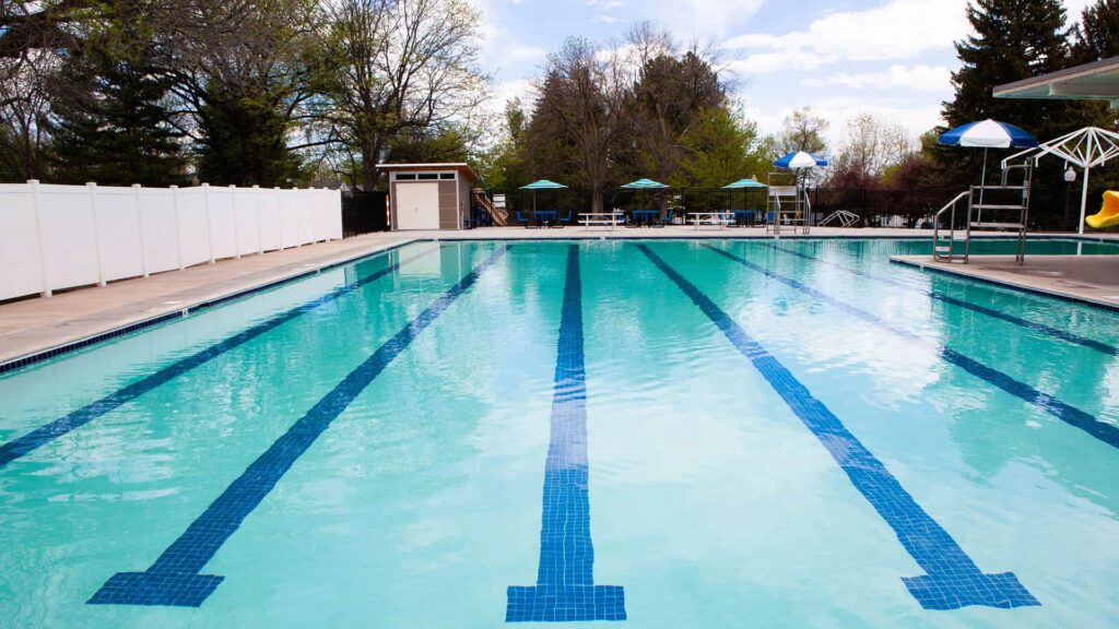 Outdoor swimming pool with clear blue water and lane markings. Surrounding are trees and loungers with blue umbrellas. The scene is serene and inviting.