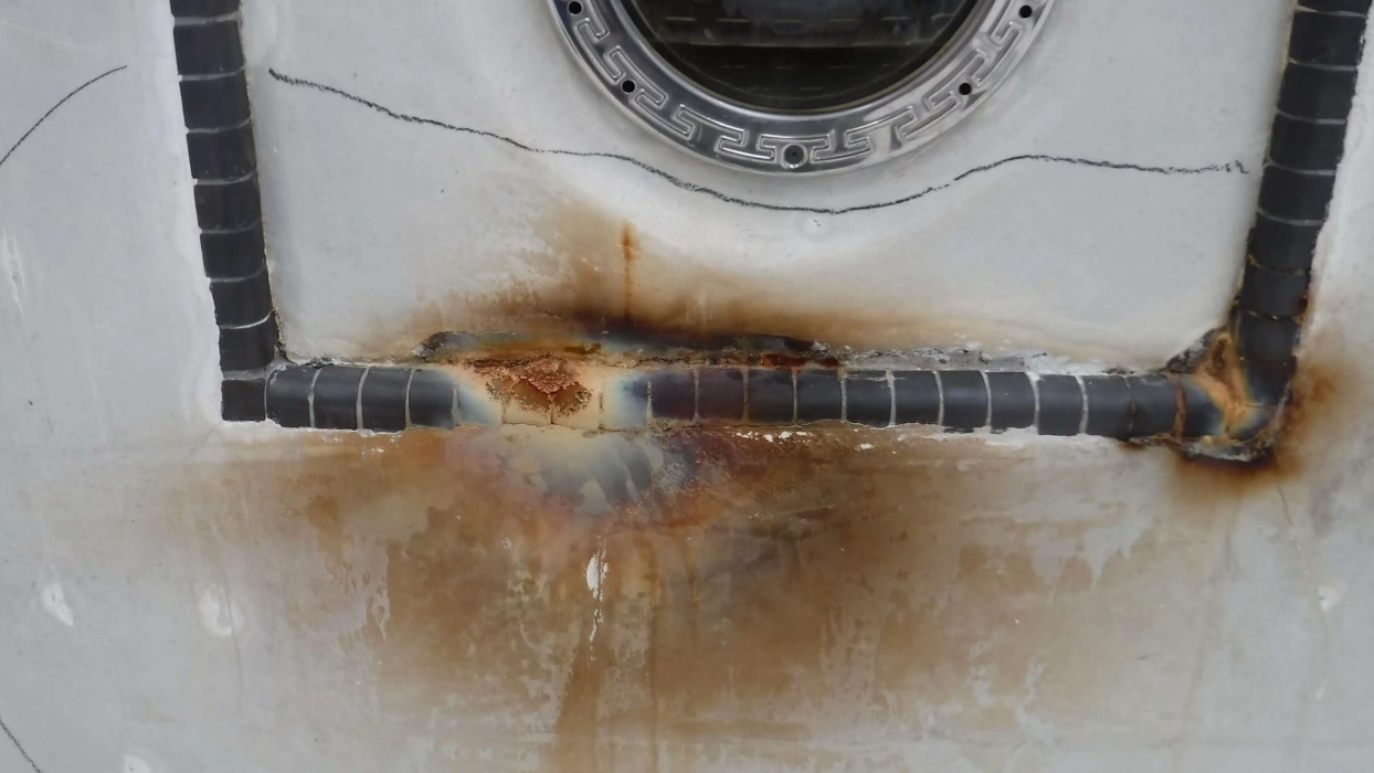 Close-up of a metal surface with significant rust damage around welded joints and a circular opening. The scene conveys deterioration and wear.