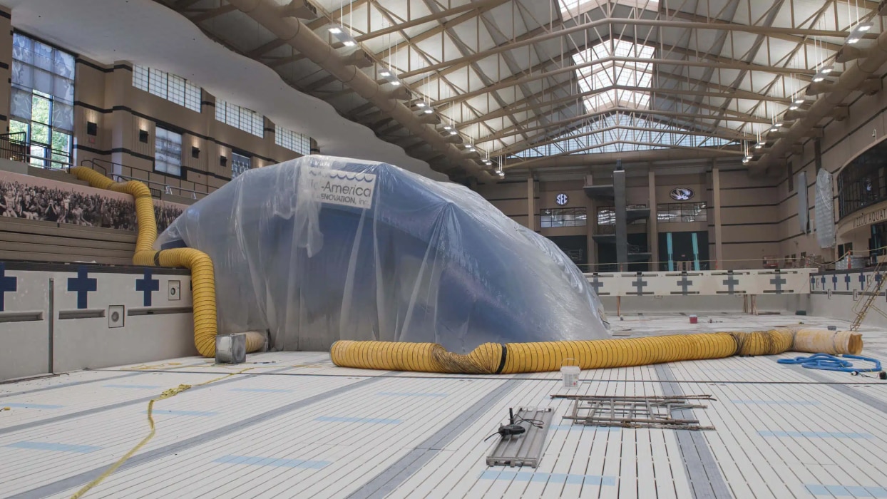 An empty indoor pool covered partially by a large plastic sheet and yellow hoses. The space is lit by natural light through a glass ceiling, creating an industrial feel.