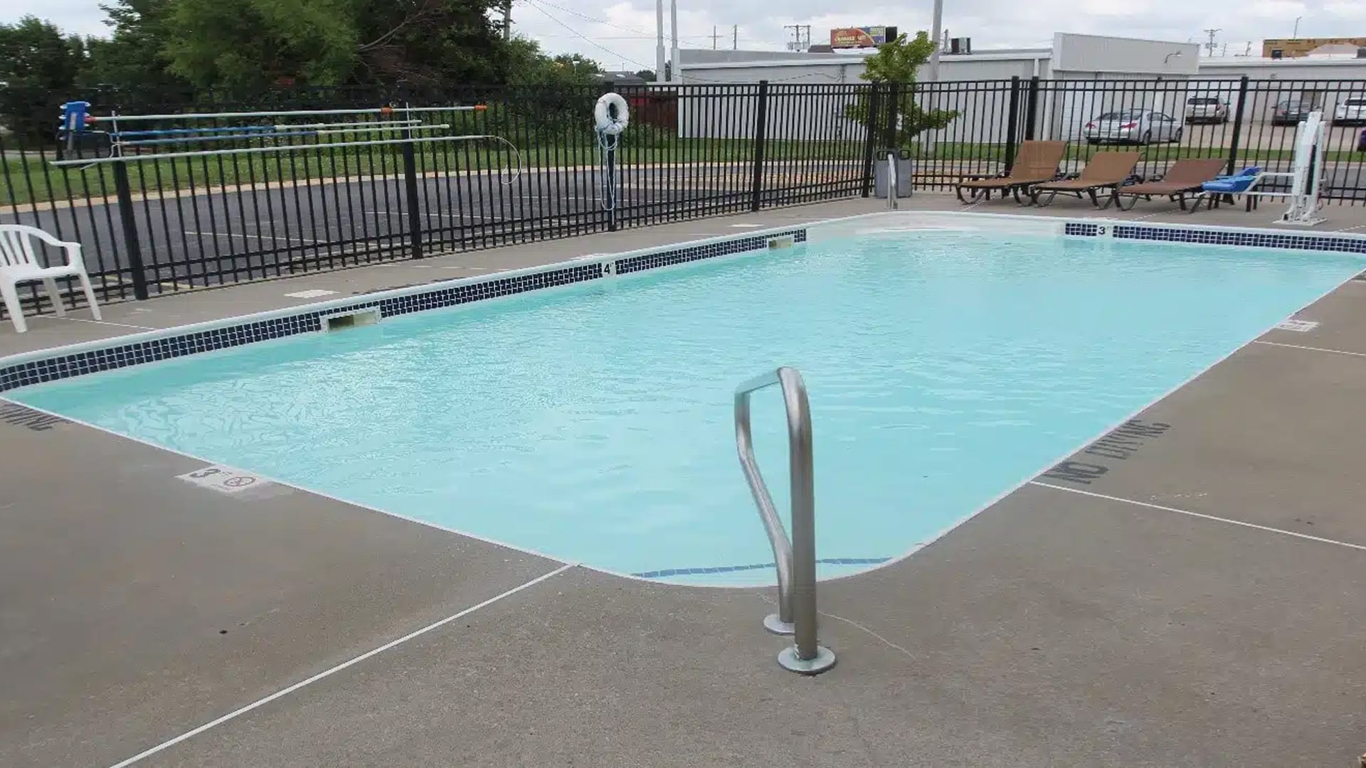 Rectangular outdoor swimming pool with clear water, surrounded by a concrete deck. Black metal fence encloses the area. Lounge chairs and a lifebuoy are visible.