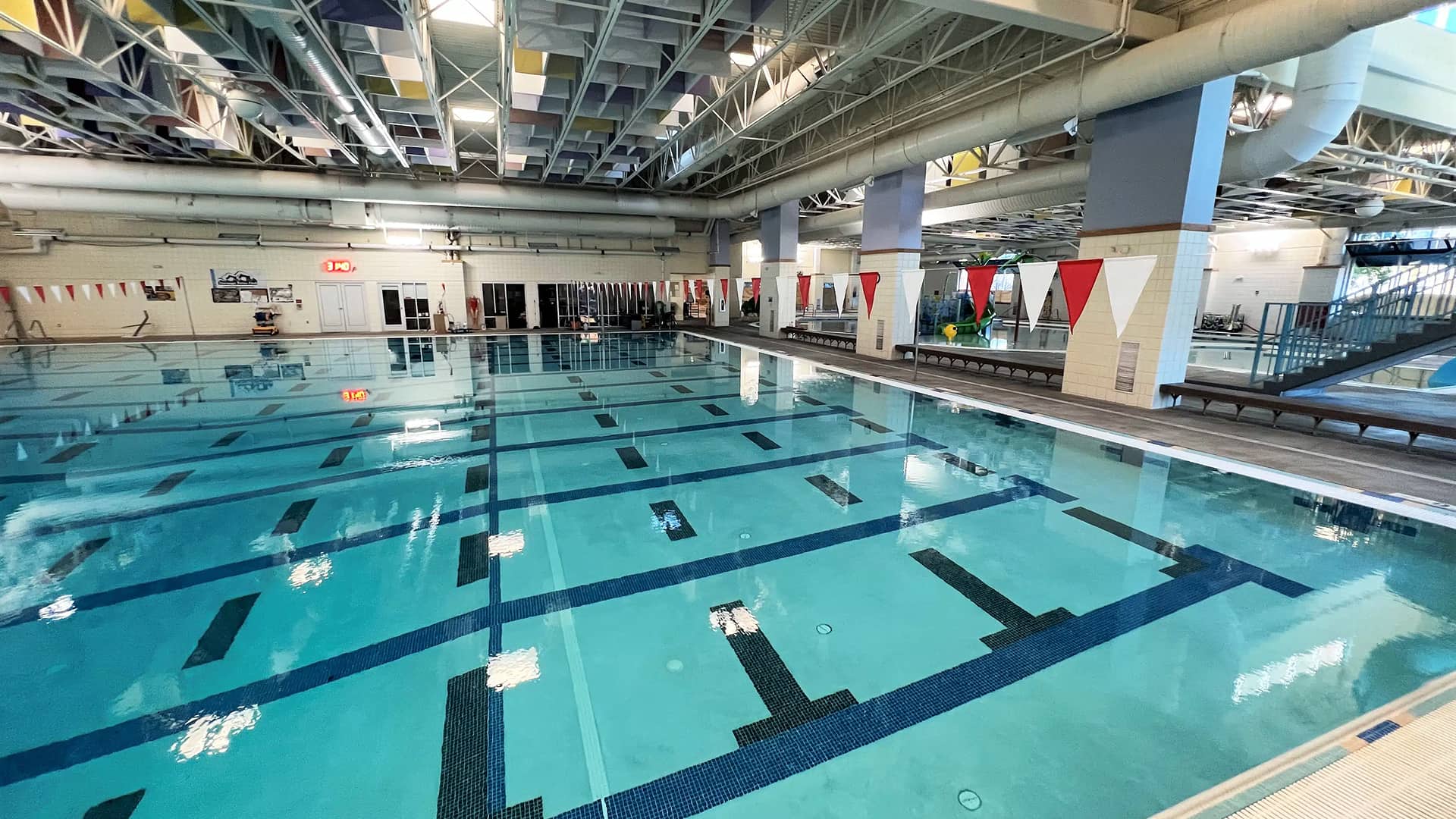 Indoor swimming pool with clear blue water and marked lanes, surrounded by a spacious, well-lit facility. Red and white flags hang above, creating a calm, inviting atmosphere.
