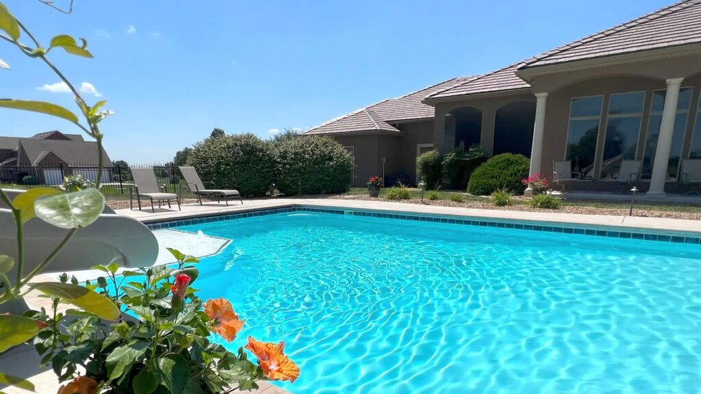 A serene backyard with a clear blue swimming pool, surrounded by lush greenery and vibrant orange flowers. Patio chairs and a modern house are in the background.