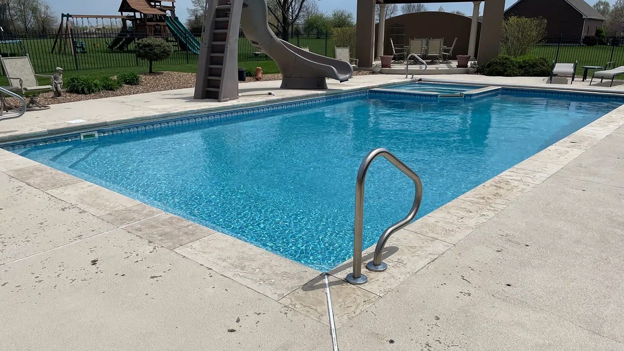 Clear blue swimming pool with metal handrails, surrounded by a concrete deck. A slide enters the pool. Playground and green lawn visible in the background. Calm and inviting atmosphere.