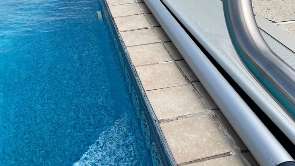 Close-up of a swimming pool's edge with clear blue water and beige tiles. A metal handrail is visible, creating a calm and inviting atmosphere.