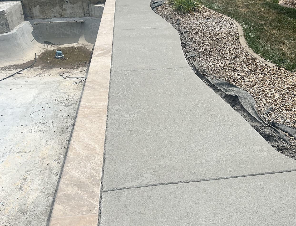 A partially constructed pool with concrete flooring, adjacent to a sidewalk. The area is unfinished, surrounded by grass and a stone-lined edge.