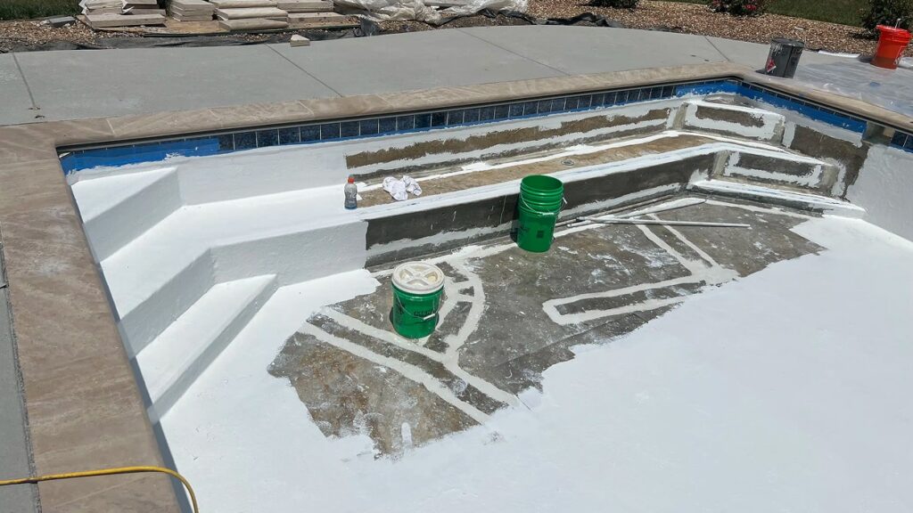 Partially completed swimming pool renovation, showing white plaster coating, exposed areas, two green buckets, and construction supplies on a sunny day.