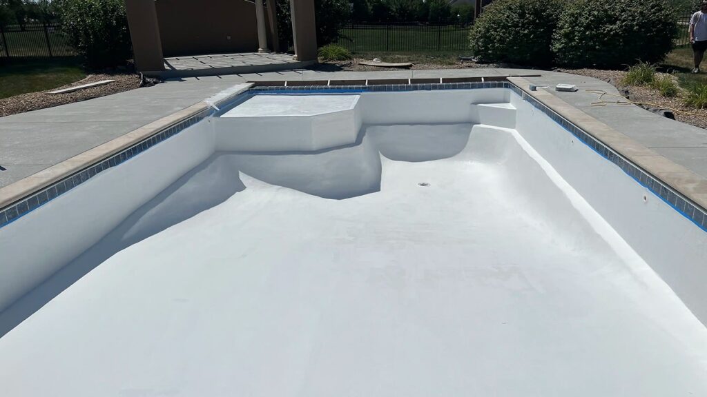 Empty swimming pool with smooth, white plastered surface and surrounding stone patio. Background includes shrubs and a partially visible building.