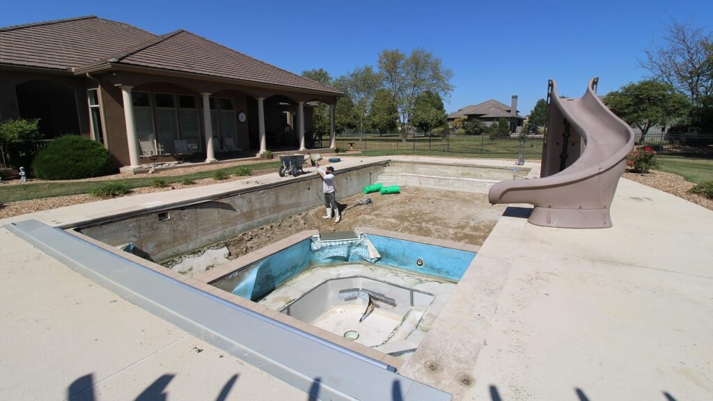 A person stands in an empty, partially demolished pool beside a house. The scene suggests renovation, with scattered debris and a slide, under a clear sky.