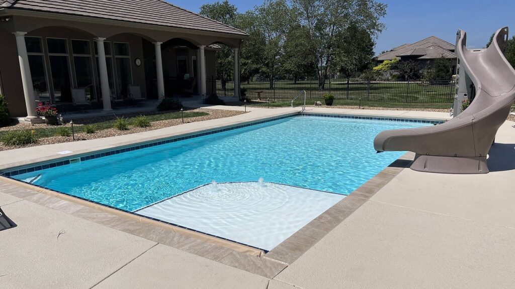 Outdoor pool with a stone patio, slide, and a house with arched windows in the background. Surrounded by greenery, it conveys a serene, sunny ambiance.