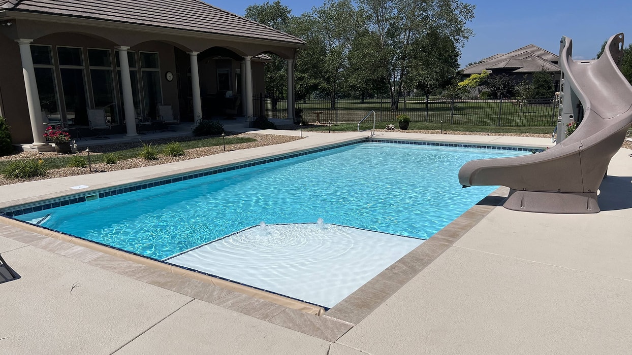 Outdoor pool with a stone patio, slide, and a house with arched windows in the background. Surrounded by greenery, it conveys a serene, sunny ambiance.